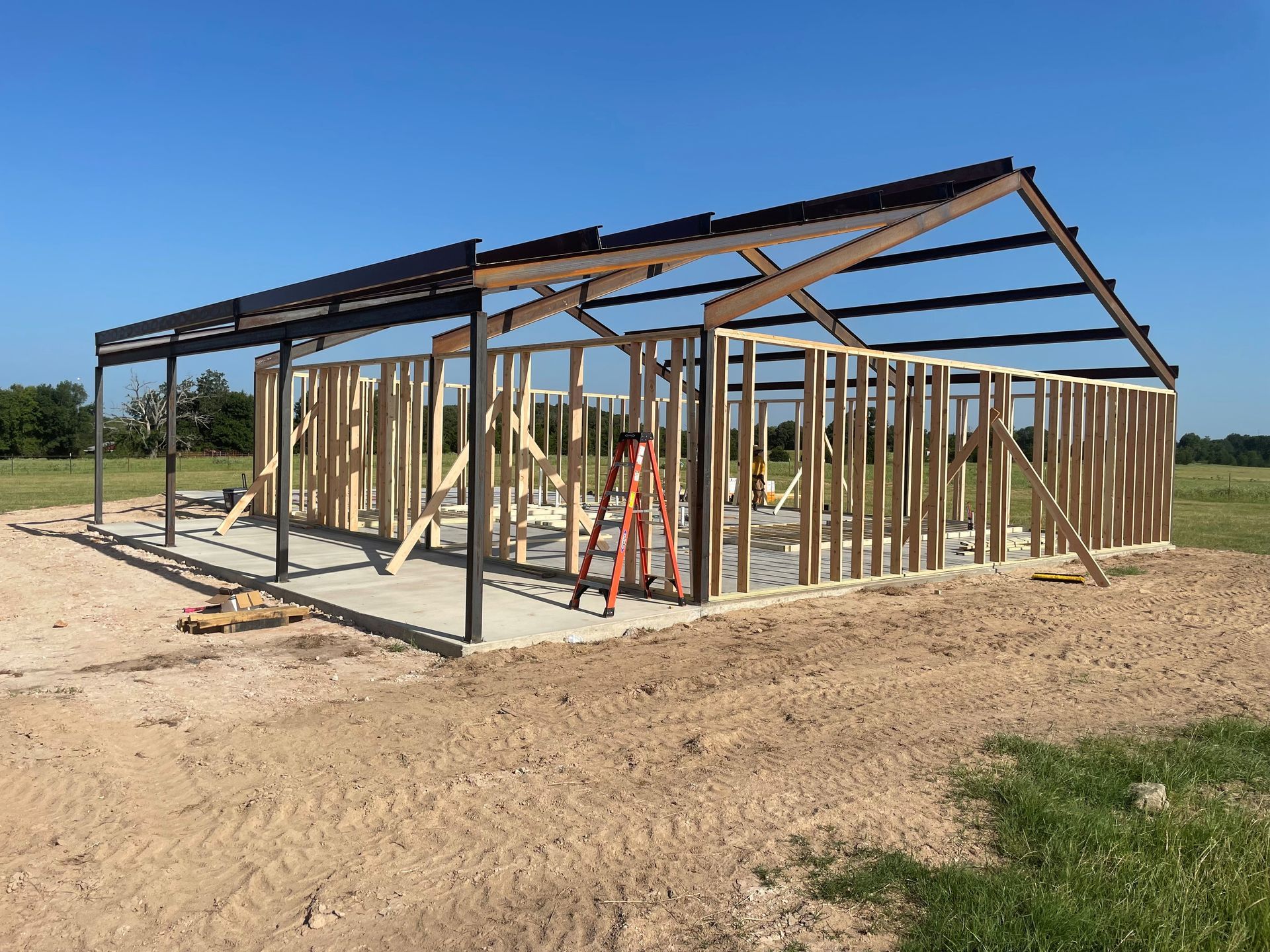 Wood-framed house under construction on a dirt lot, with a blue sky and metal roof beams.