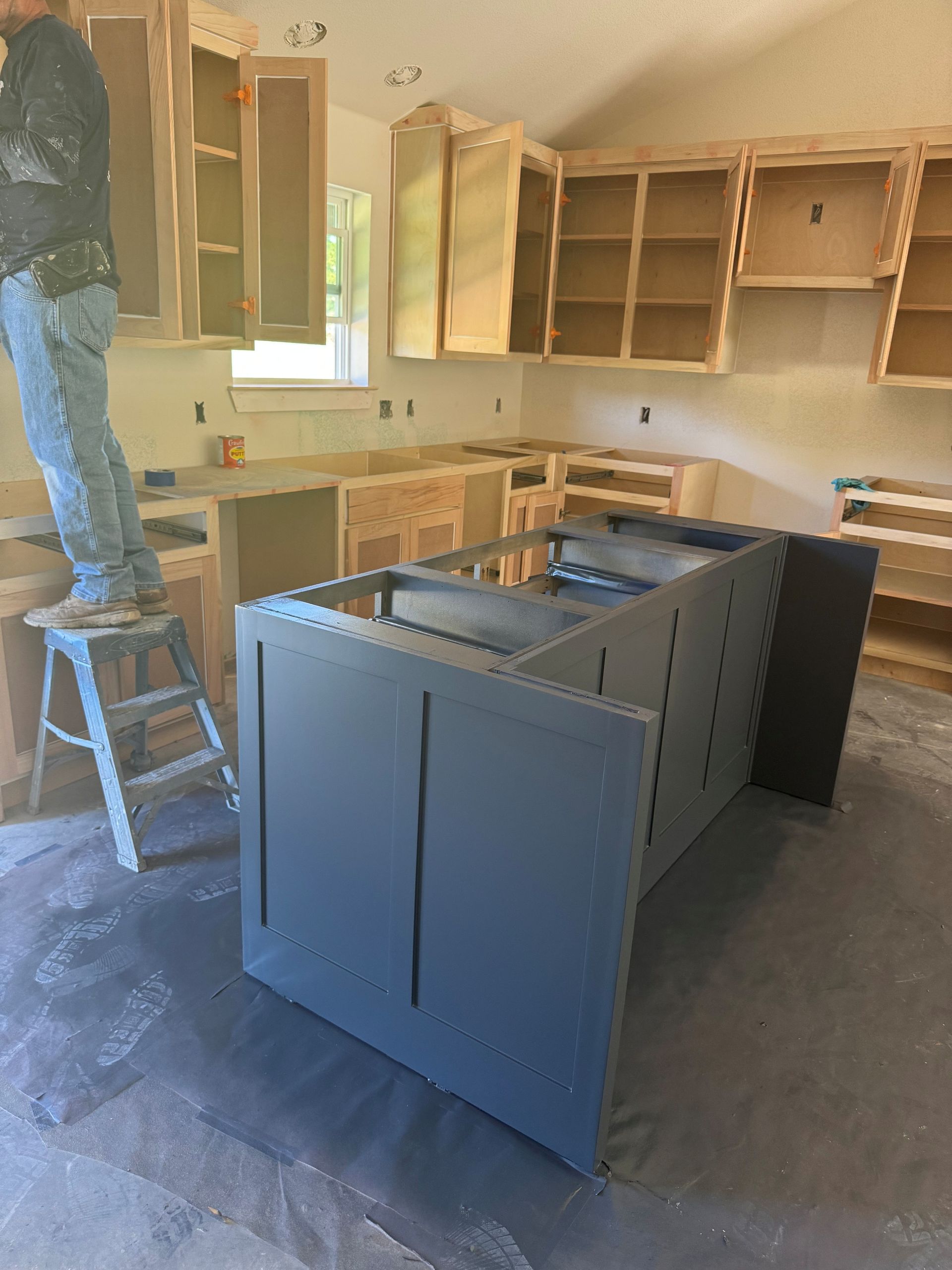 Unfinished kitchen with blue island cabinets, tan wall cabinets, and a worker on a ladder installing upper cabinets