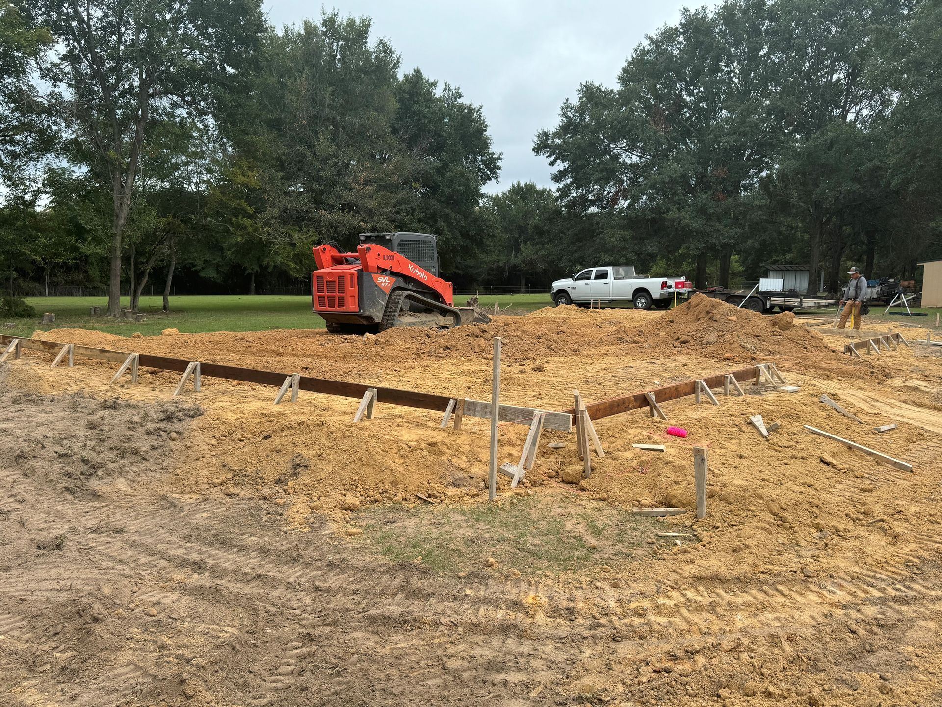Construction site with orange skid steer, dirt lot, and white pickup truck near trees