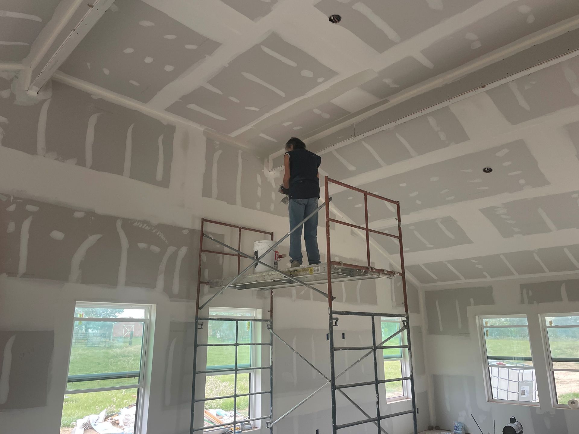 Worker on scaffolding finishing drywall in a room with tall ceilings and windows