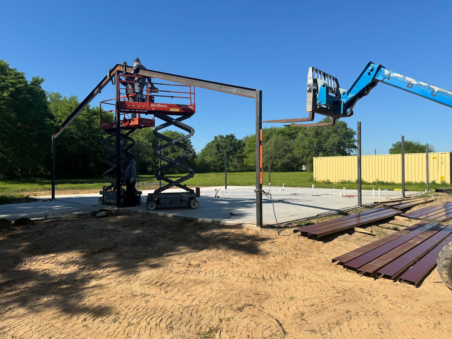 Construction lift and boom arm beside a steel frame on a sandy worksite with stacked metal beams.