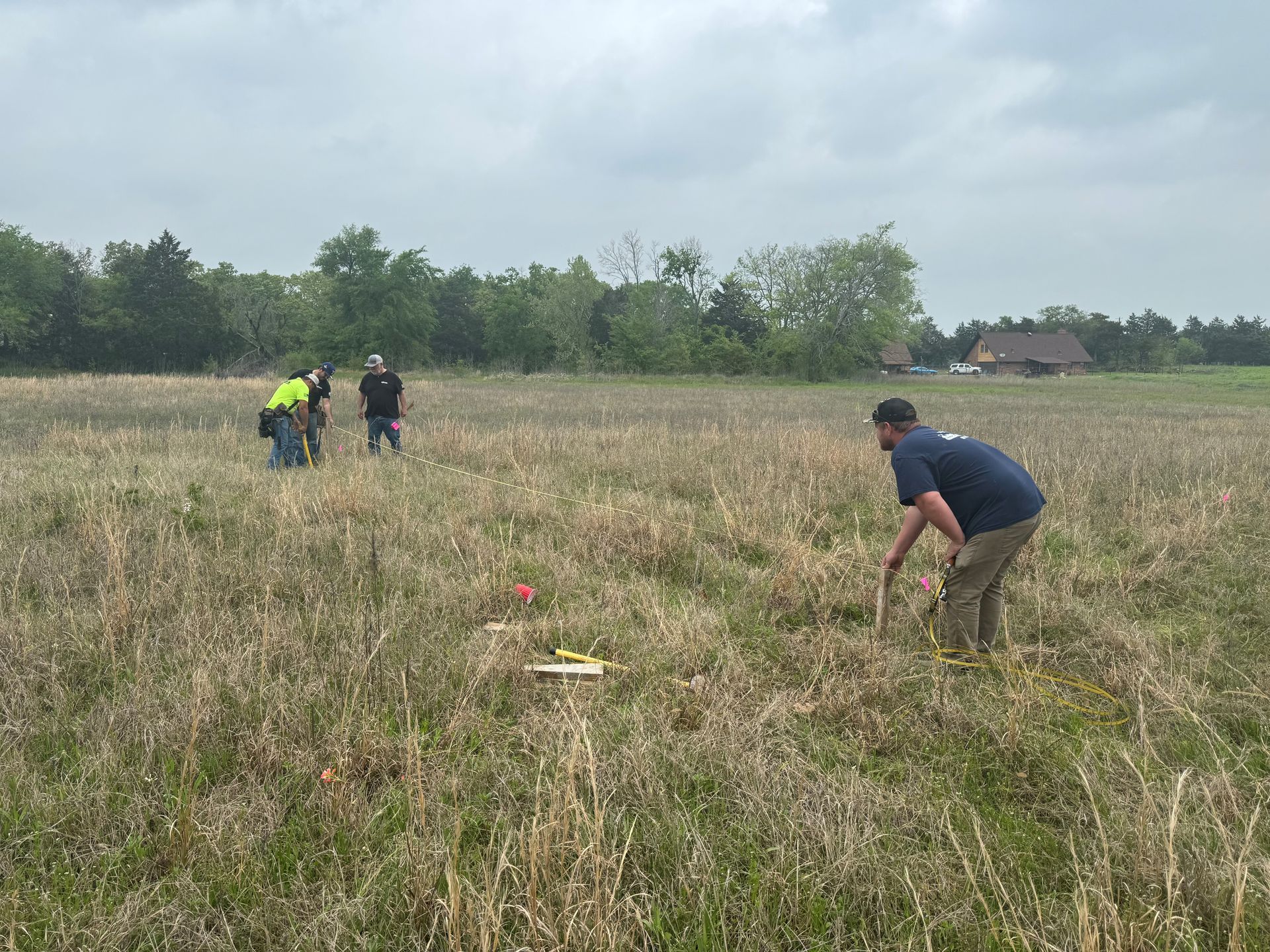 People in a dry field marking spots, with one person crouching in the foreground and trees in the background