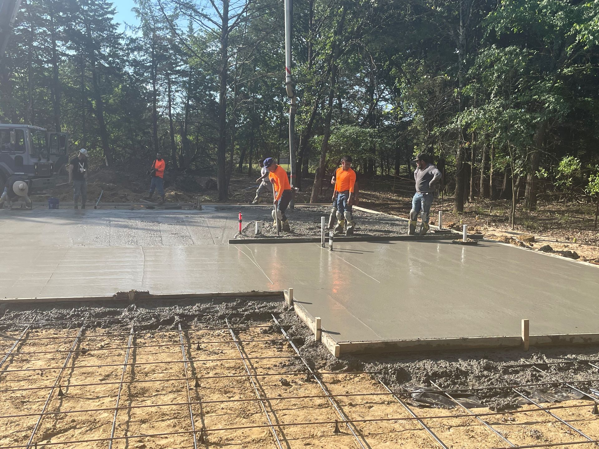 Workers pouring concrete at an outdoor construction site with muddy slabs and a wooded background