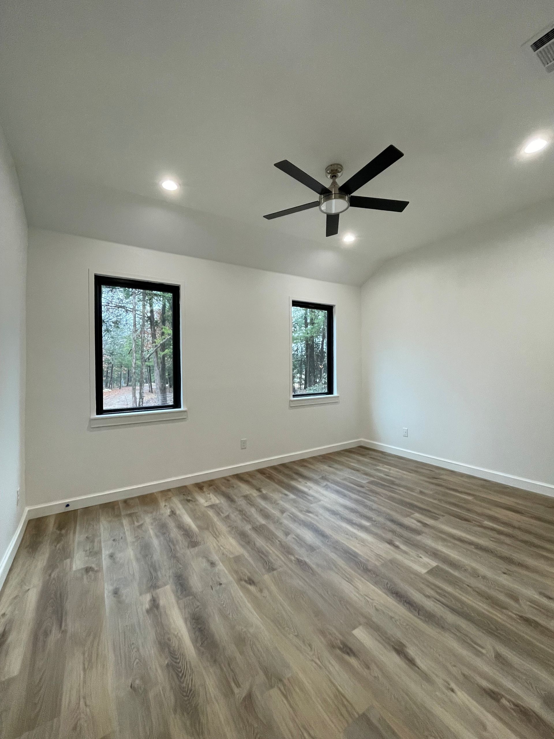 Empty modern room with wood floors, white walls, two windows, and a black ceiling fan.