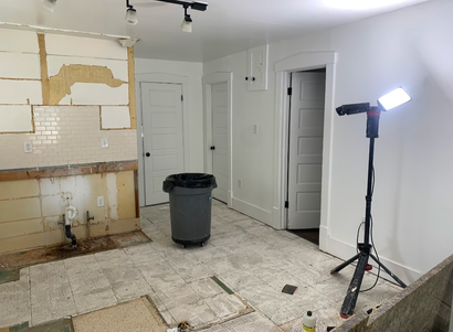 Renovated kitchen with exposed wall repairs, tiled floor, trash bin, and work light on tripod
