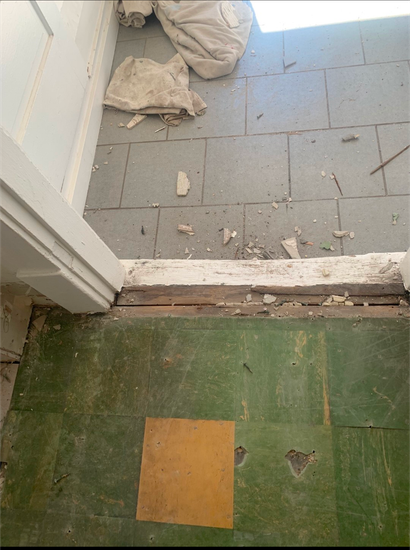 Green and gray tiled floor with a damaged threshold and debris near stacked bags