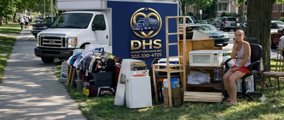 A woman sits in a chair next to a collection of household items and a Lowry Place moving truck parked on a grass verge.