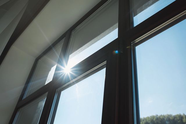 Il sole splendente filtra attraverso un'ampia finestra con infissi scuri, proiettando un bagliore che si riflette sul cielo azzurro e limpido.