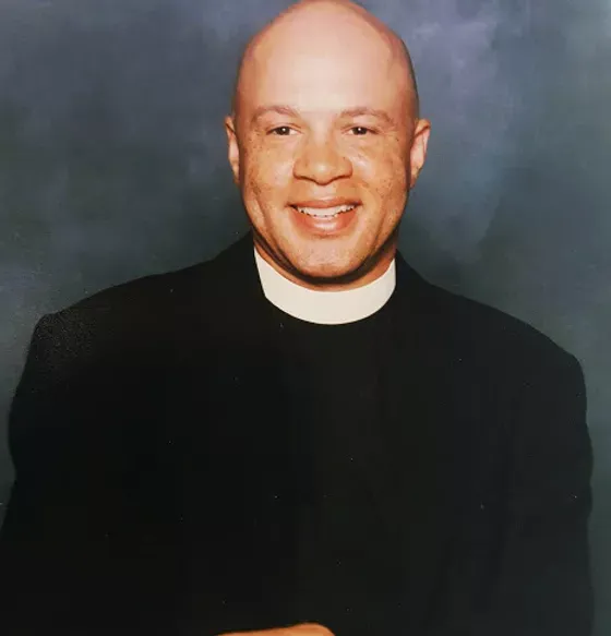 A man in a black suit and white collar is smiling.