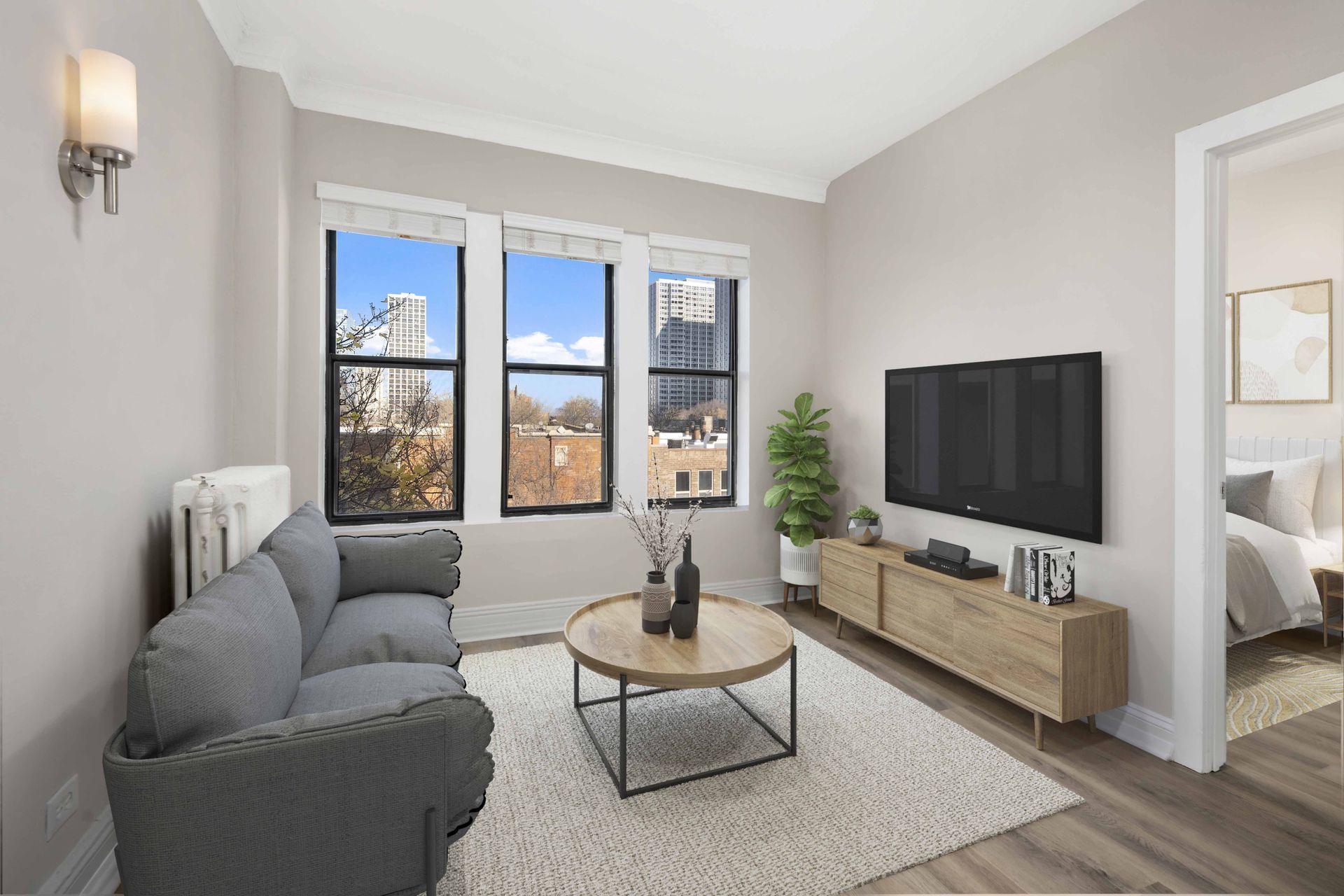 A living room with a grey couch, a coffee table, a flat screen TV and a window with a view of the city.