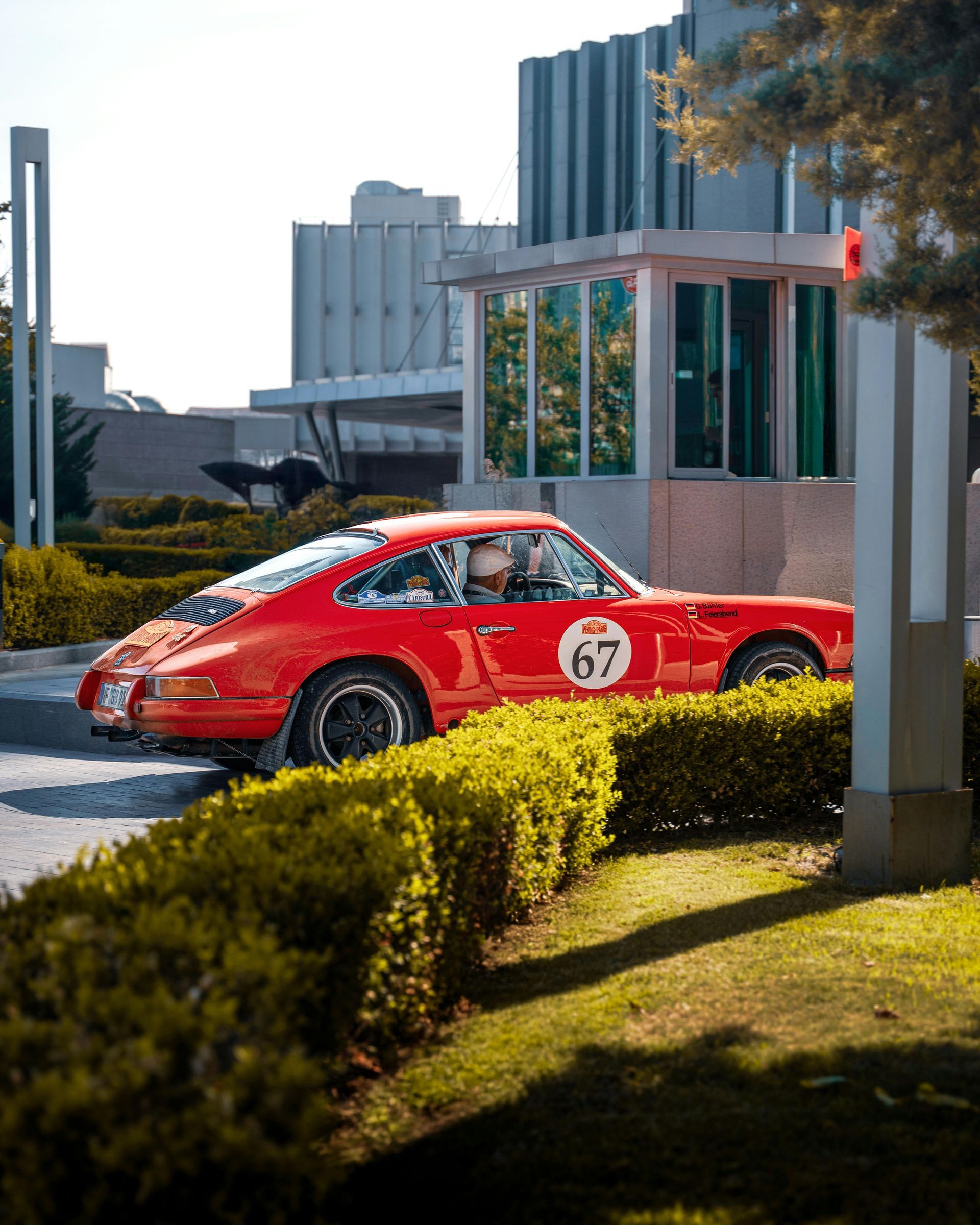 Red classic Porsche 911 with race number 67 driving near a building entrance, sunny day.