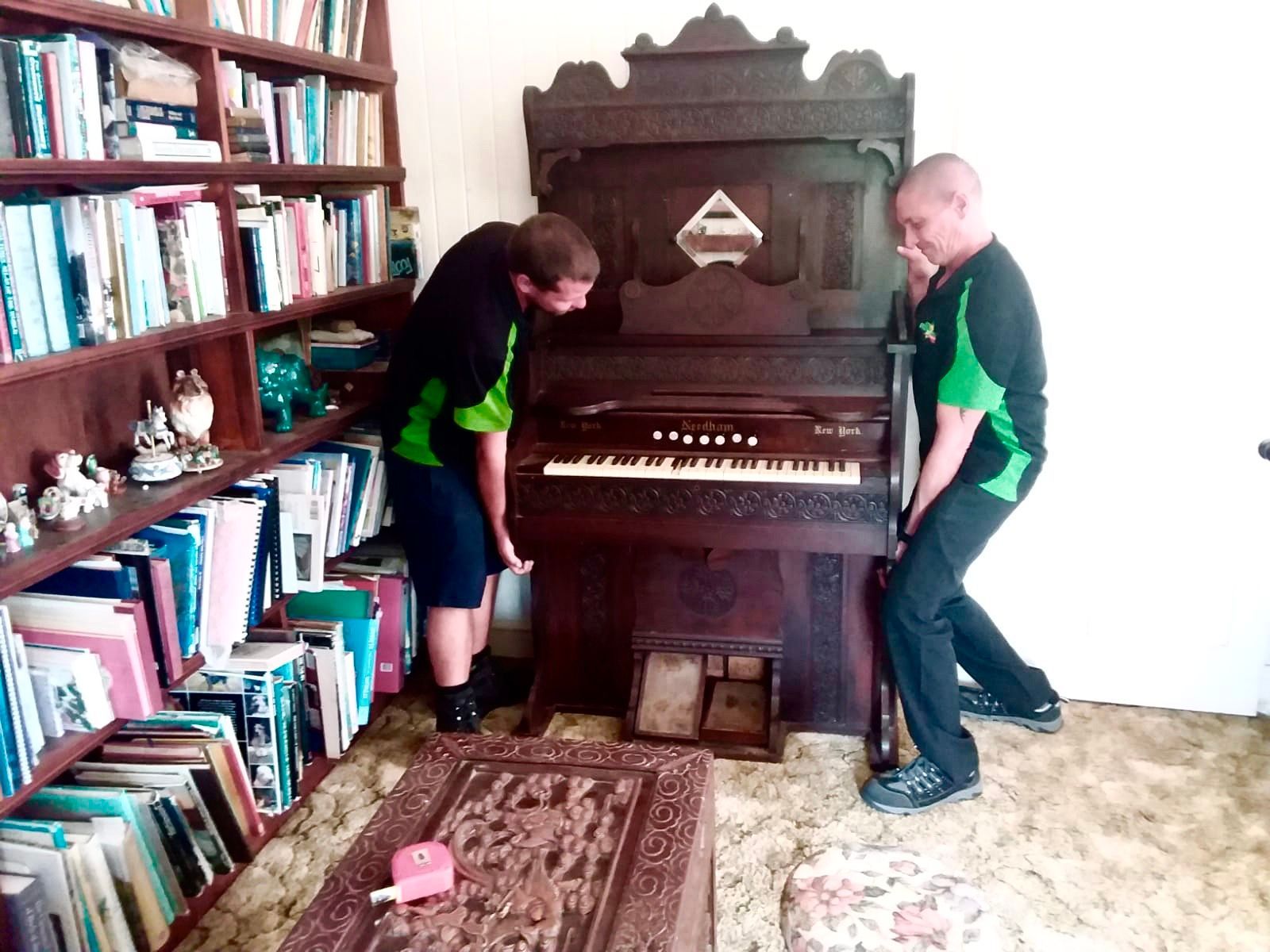 Two People Lifting a Dark Wooden Organ in a Room With a Bookshelf and Carpet — Little Green Truck Bundaberg In Hervey Bay, QLD