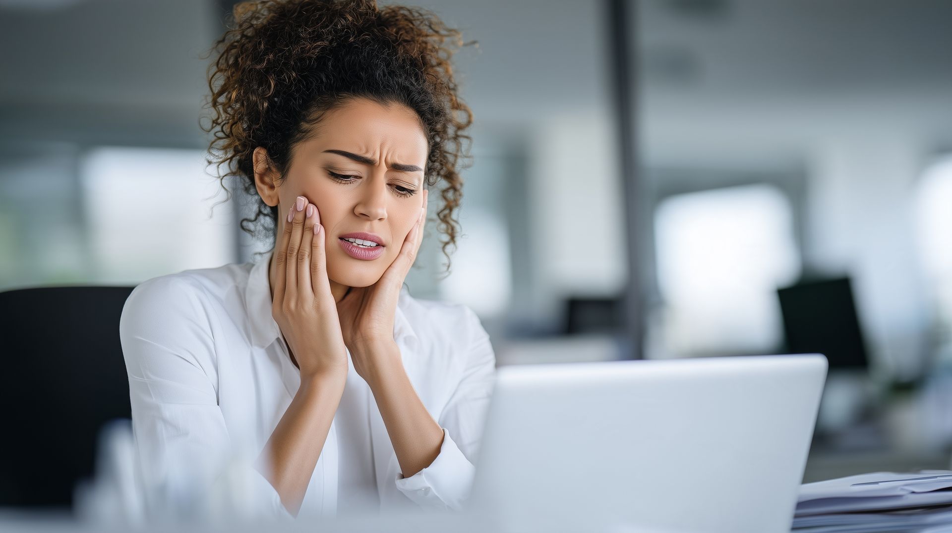 Woman wincing and holding her cheek at her desk, in need of an emergency dentist