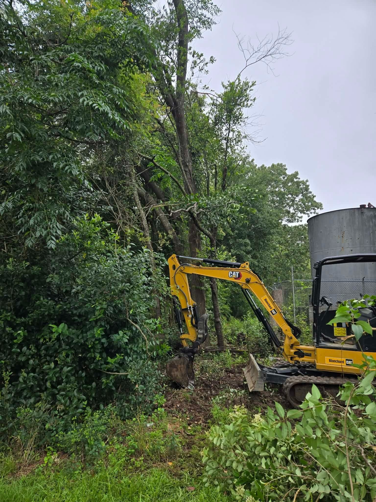 Yellow excavator clearing dense vegetation beside a large gray tank in a wooded area