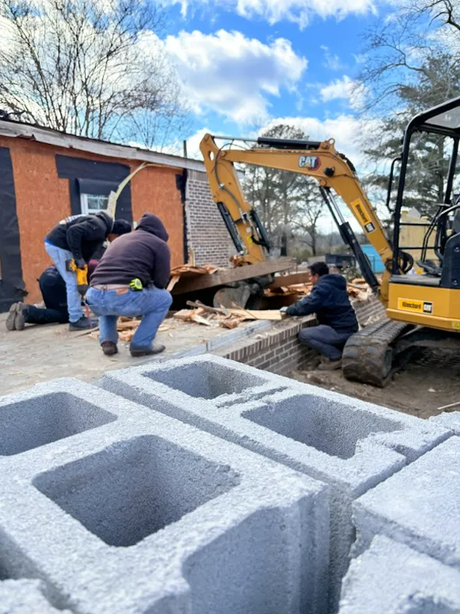 Workers use a yellow excavator to move debris beside stacked gray cinder blocks at a construction site