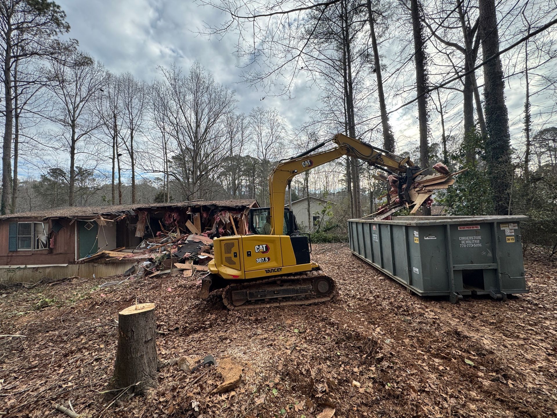 Yellow excavator clearing brush beside a green dumpster in a wooded yard near a house