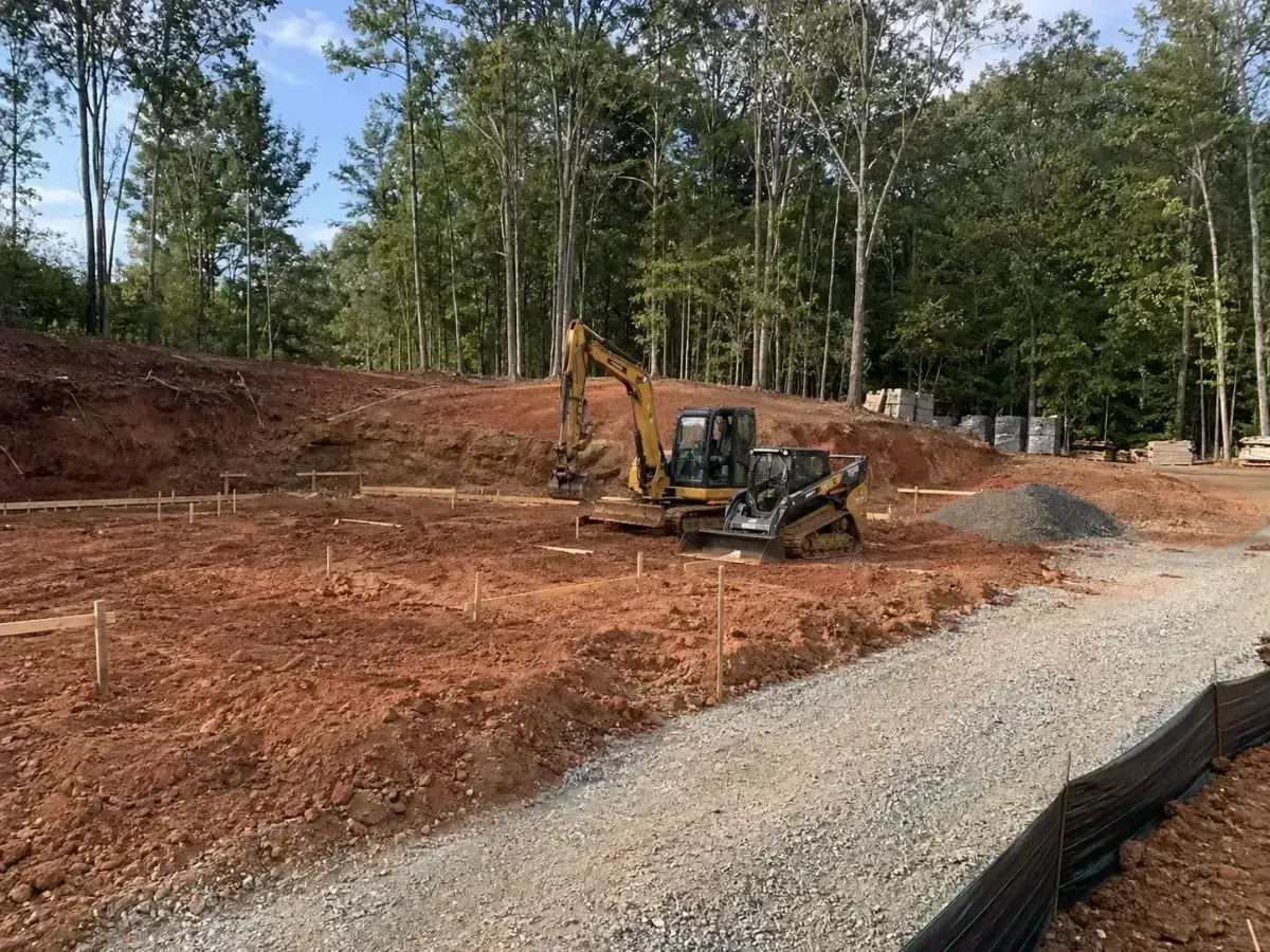 Excavator grading red dirt on a construction site beside a gravel road and trees