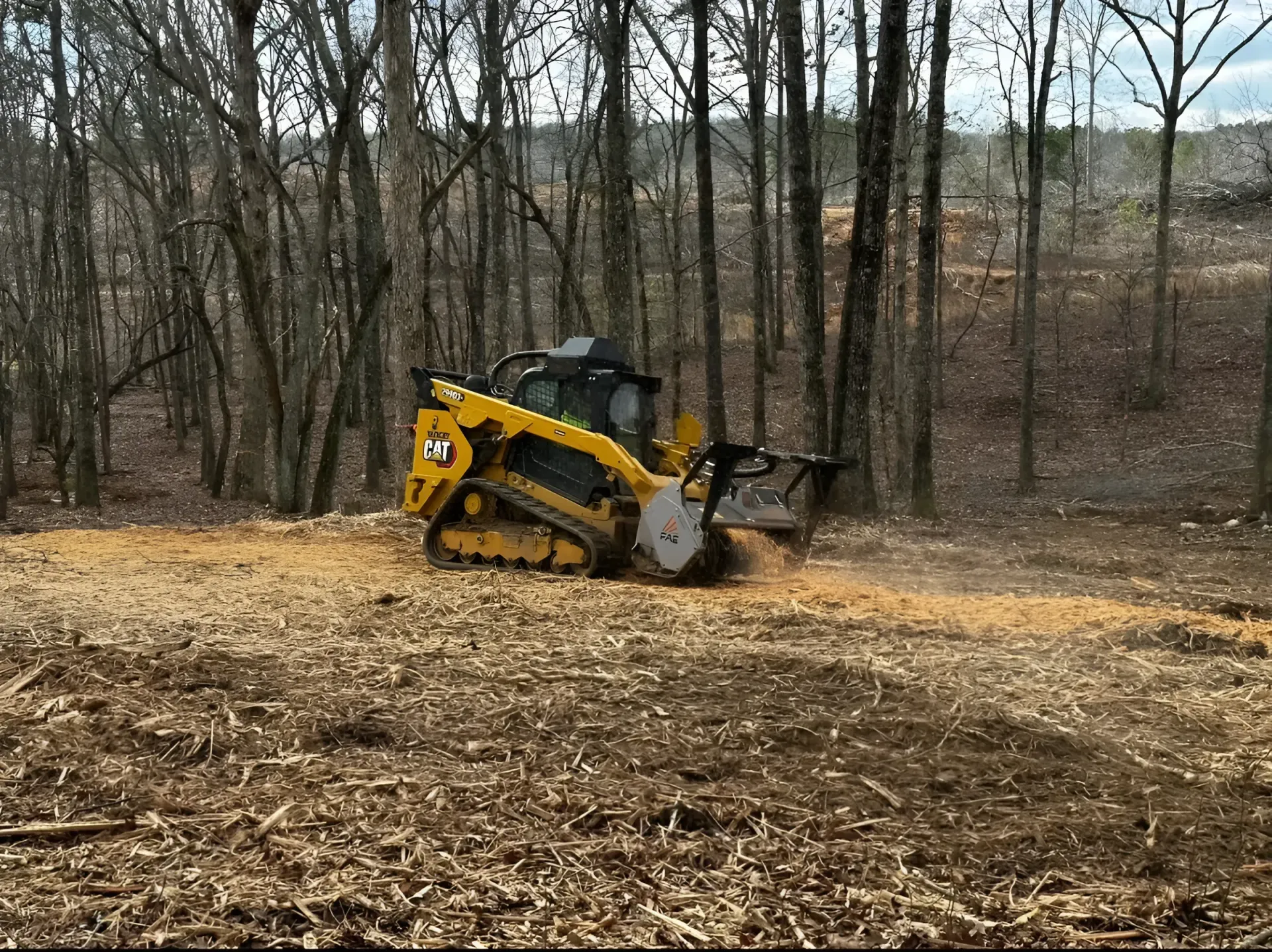 Yellow skid steer clearing muddy ground in a wooded area with bare trees
