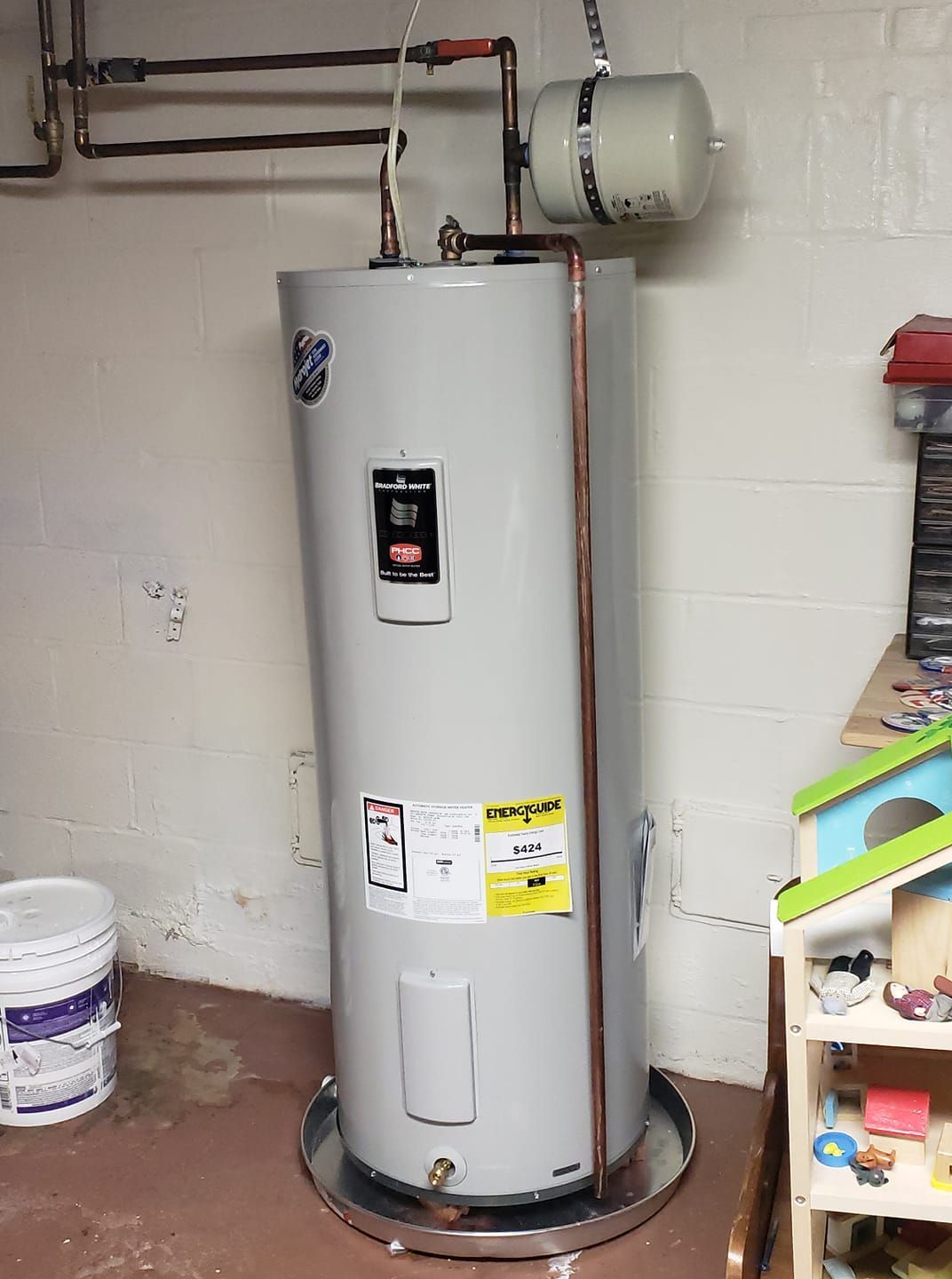 A water heater is sitting in a basement next to a toy house.