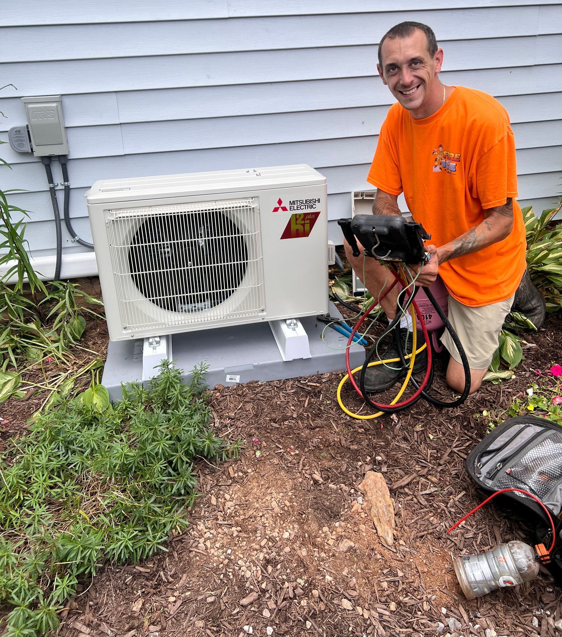 A man in an orange shirt is kneeling in front of an air conditioner.