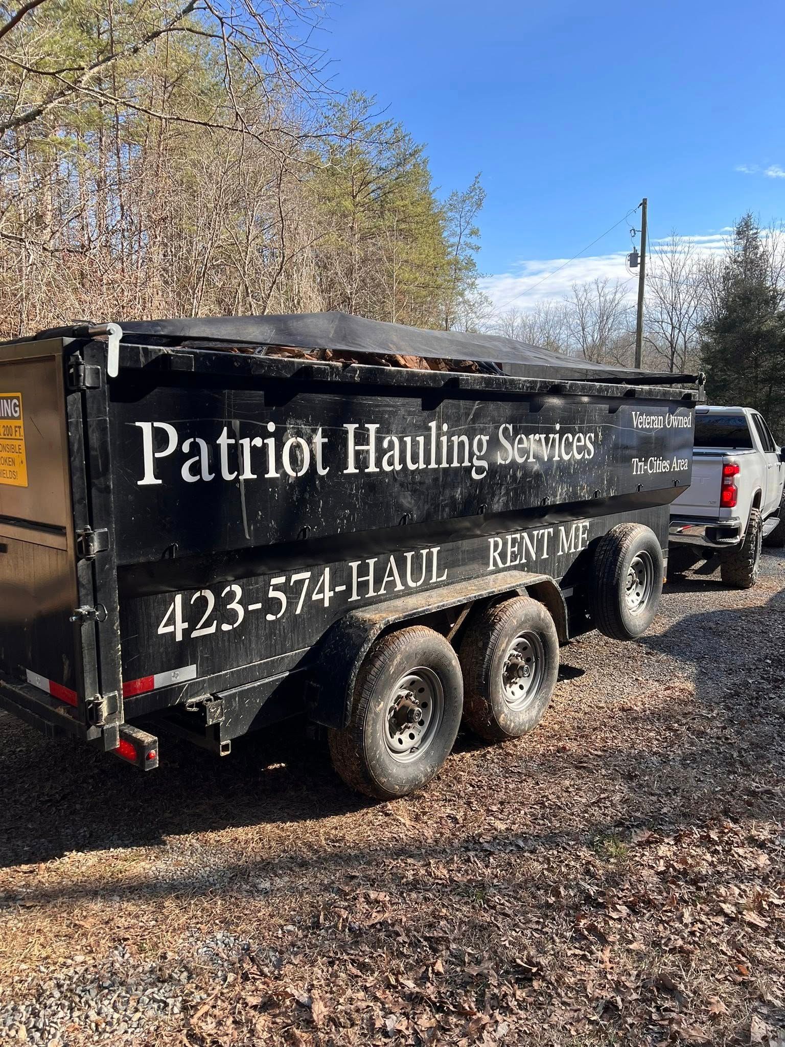 Black Patriot Hauling Services dump trailer parked on gravel with trees and a blue sky behind it