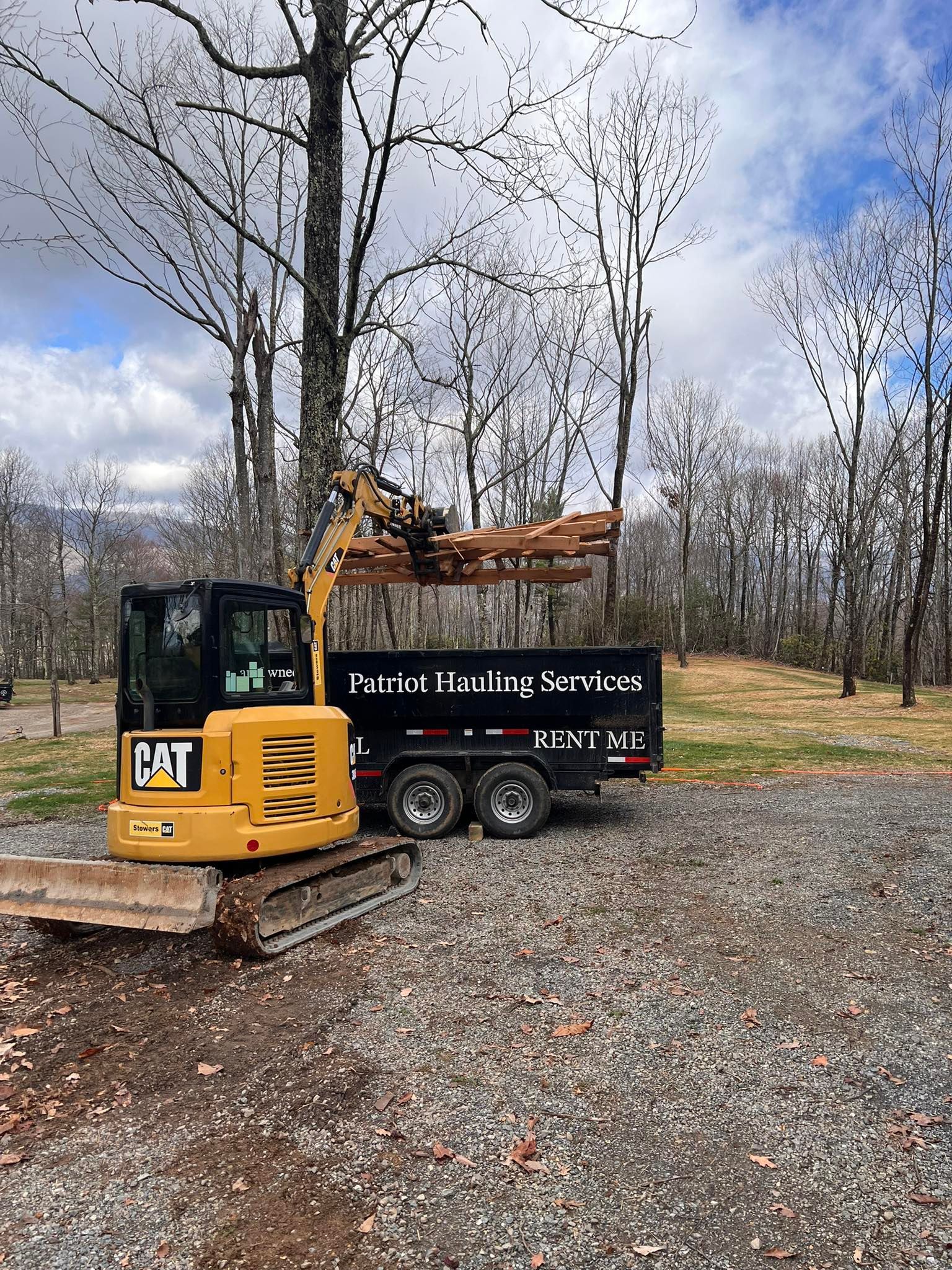 Yellow CAT excavator on a trailer in a wooded area under a cloudy sky.
