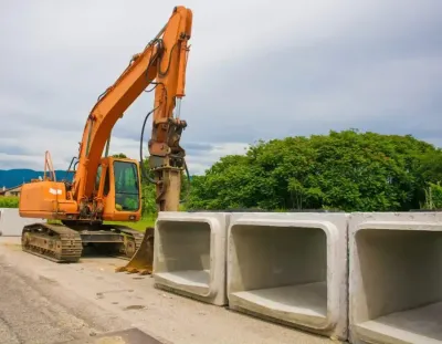 Orange excavator beside stacked concrete culverts at a construction site, with green trees in the background