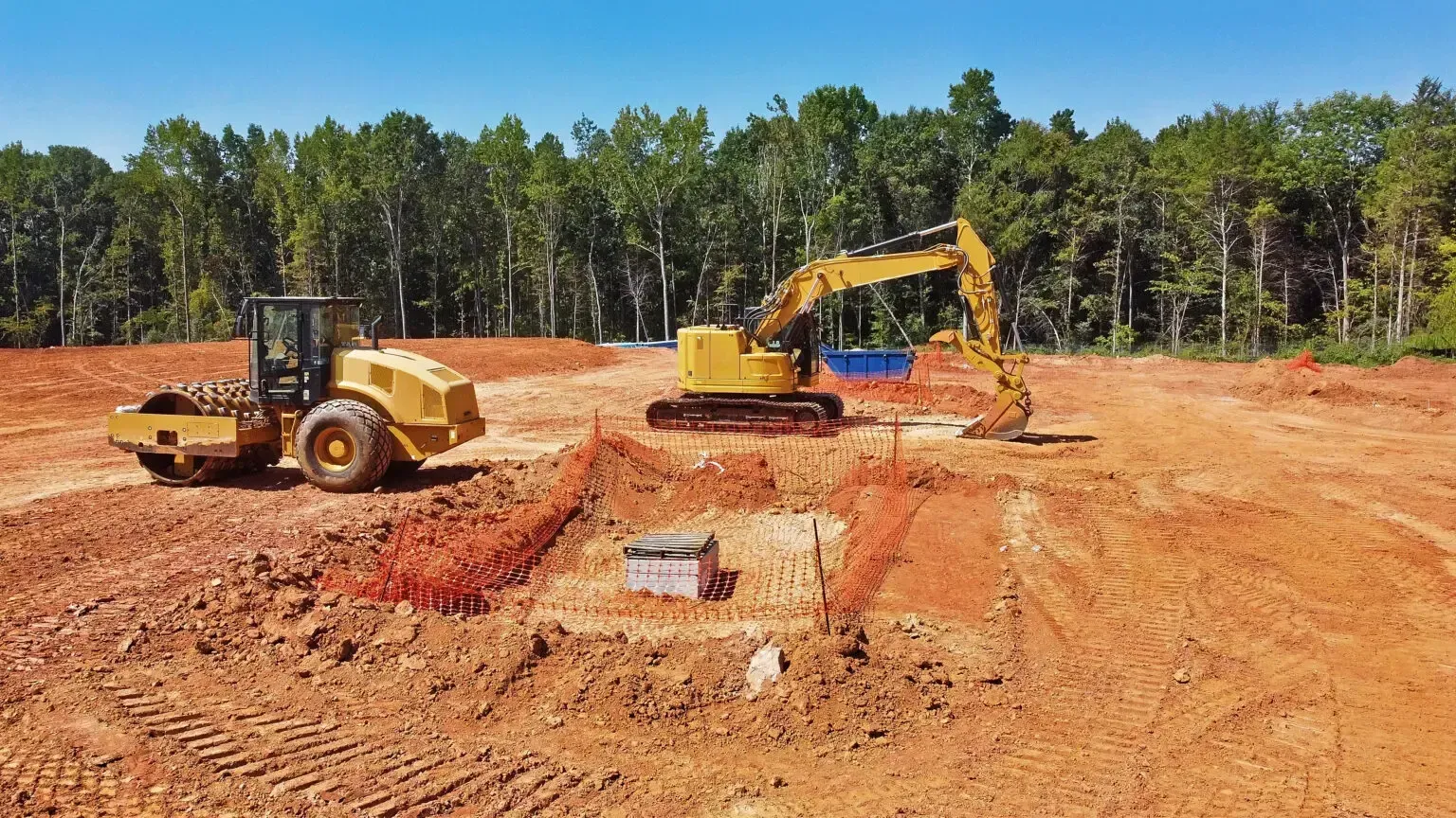 Construction site with two yellow excavators on a red dirt clearing beside a tree line