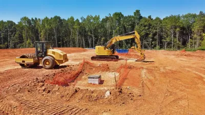 Construction site with two yellow excavators on a red dirt clearing beside a tree line