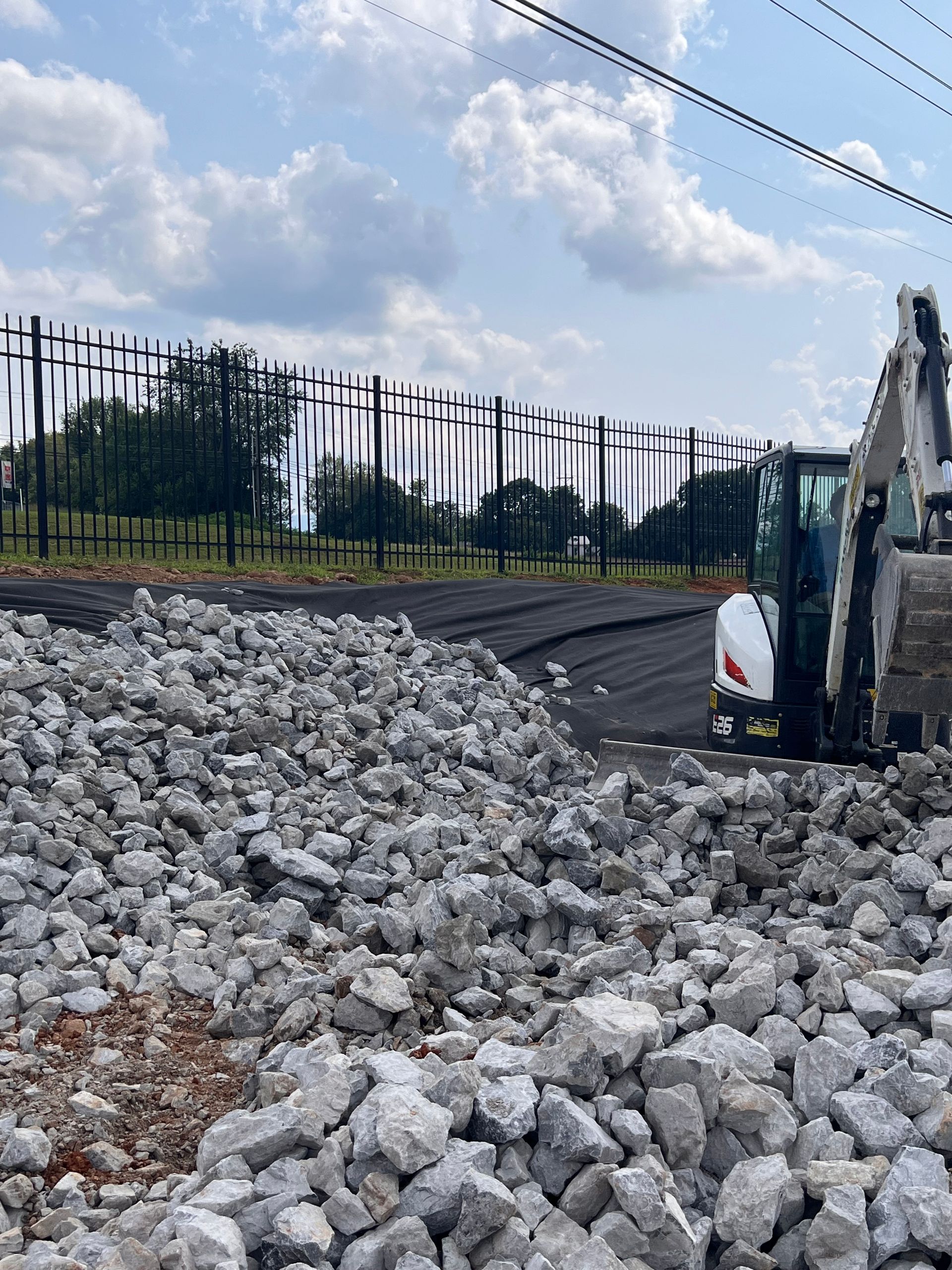 Construction site with large pile of crushed rock, excavator, and a black fence under a cloudy sky