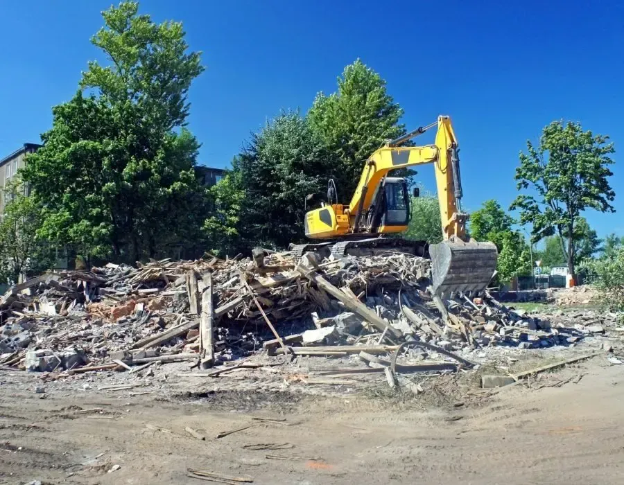 Yellow excavator demolishing a pile of rubble at a construction site with trees in the background