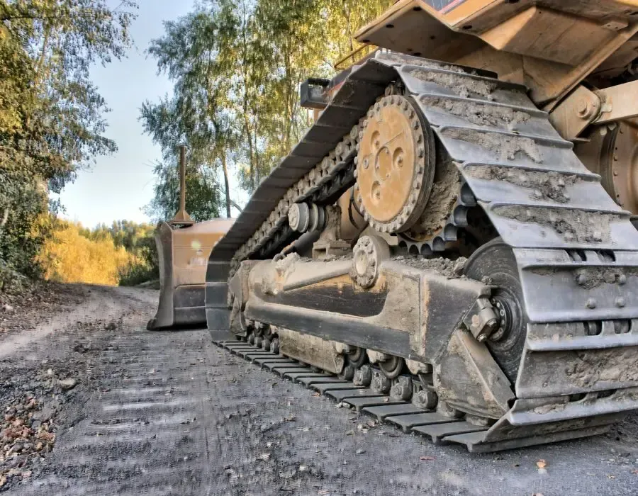 Close-up of a bulldozer track on a dirt road, with trees and sunlight in the background