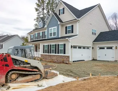 Red skid steer beside a large beige house under construction with white garage doors and unfinished yard