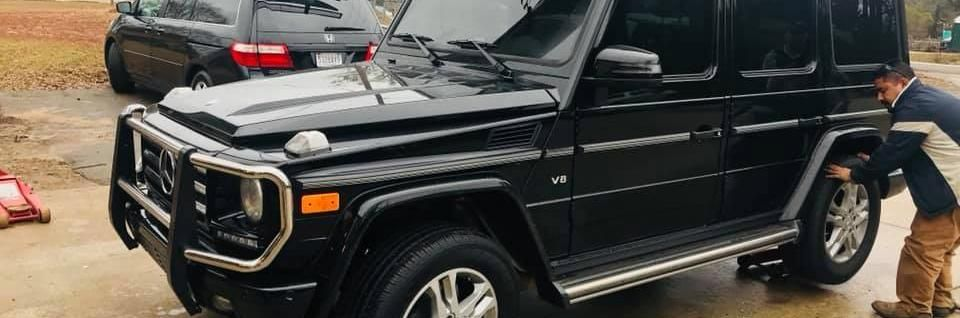 A person in work clothes uses a floor jack to lift the rear wheel of a black Mercedes-Benz G-Class SUV on a paved lot.