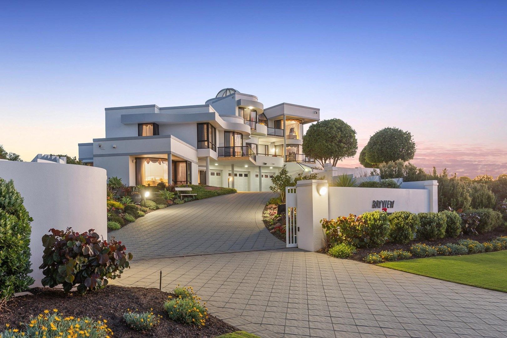 Modern white mansion with a long driveway at dusk.