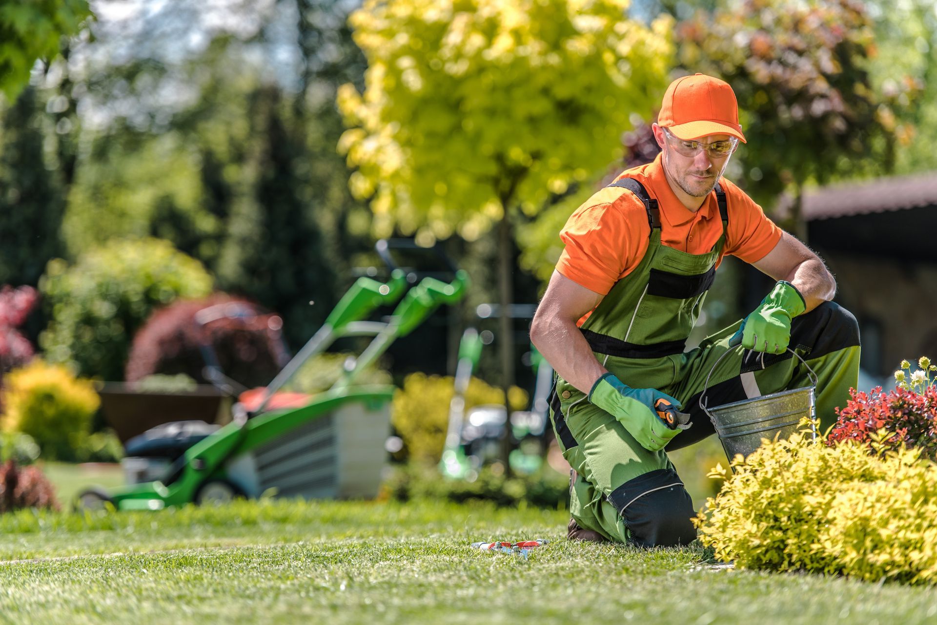 Gardener tending to a bush, kneeling in a yard with a lawnmower, wearing orange and green work clothes.