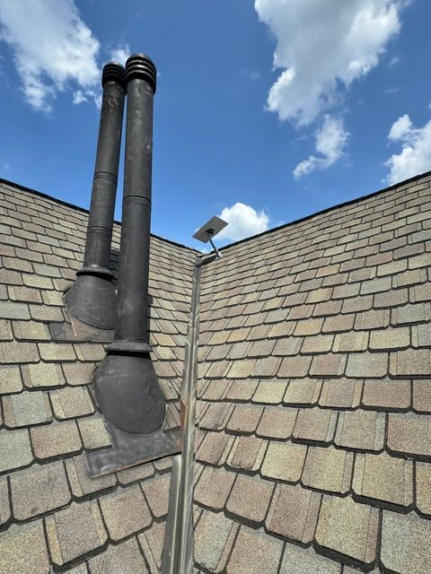 Two black chimney pipes on a gray shingled roof under a partly cloudy blue sky