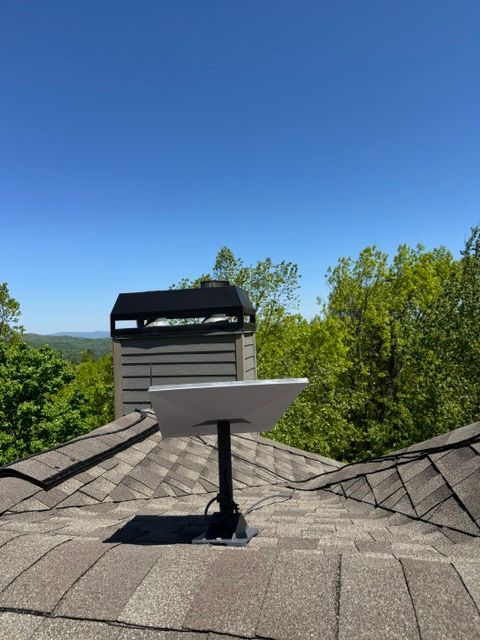 Coin-operated binocular viewer on a shingled rooftop overlooking green trees and distant hills under a clear blue sky