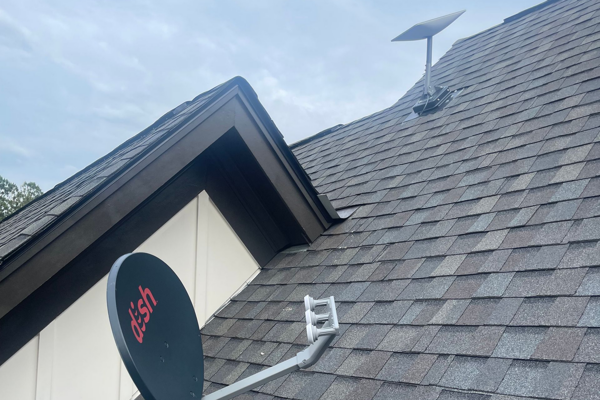 Roof with dark shingles, satellite dish, and solar panel against a blue sky.