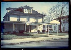 Two-story house with a porch and white awning beside a street on a sunny day