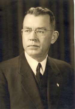 Portrait of a man in a dark suit and tie, wearing glasses, posed against a plain background