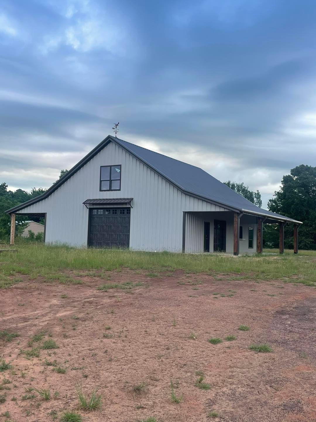 A white barn with a black garage door is sitting in the middle of a dirt field.