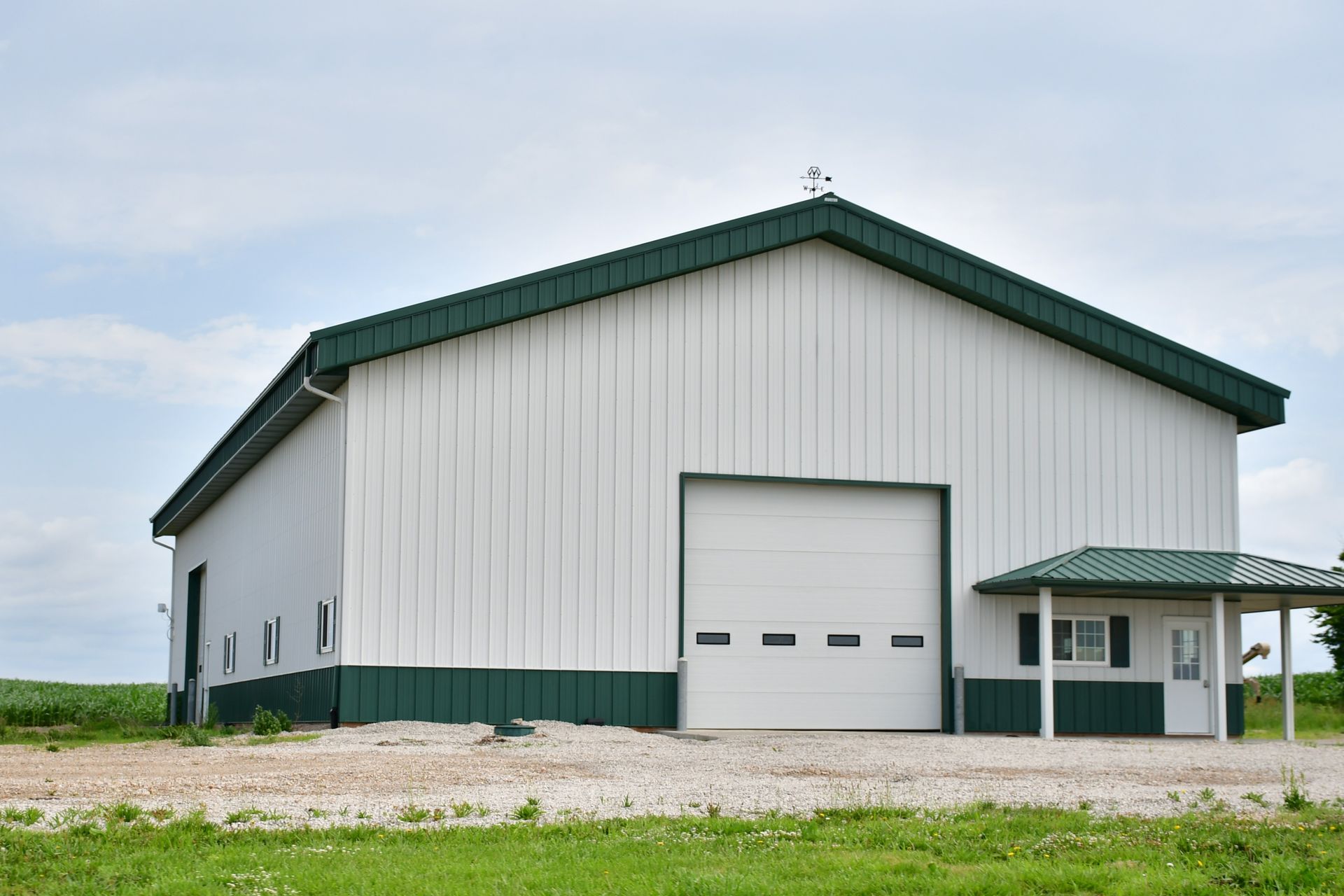 White and green barn with a large garage door and small porch, set on a gravel driveway, with blue sky.
