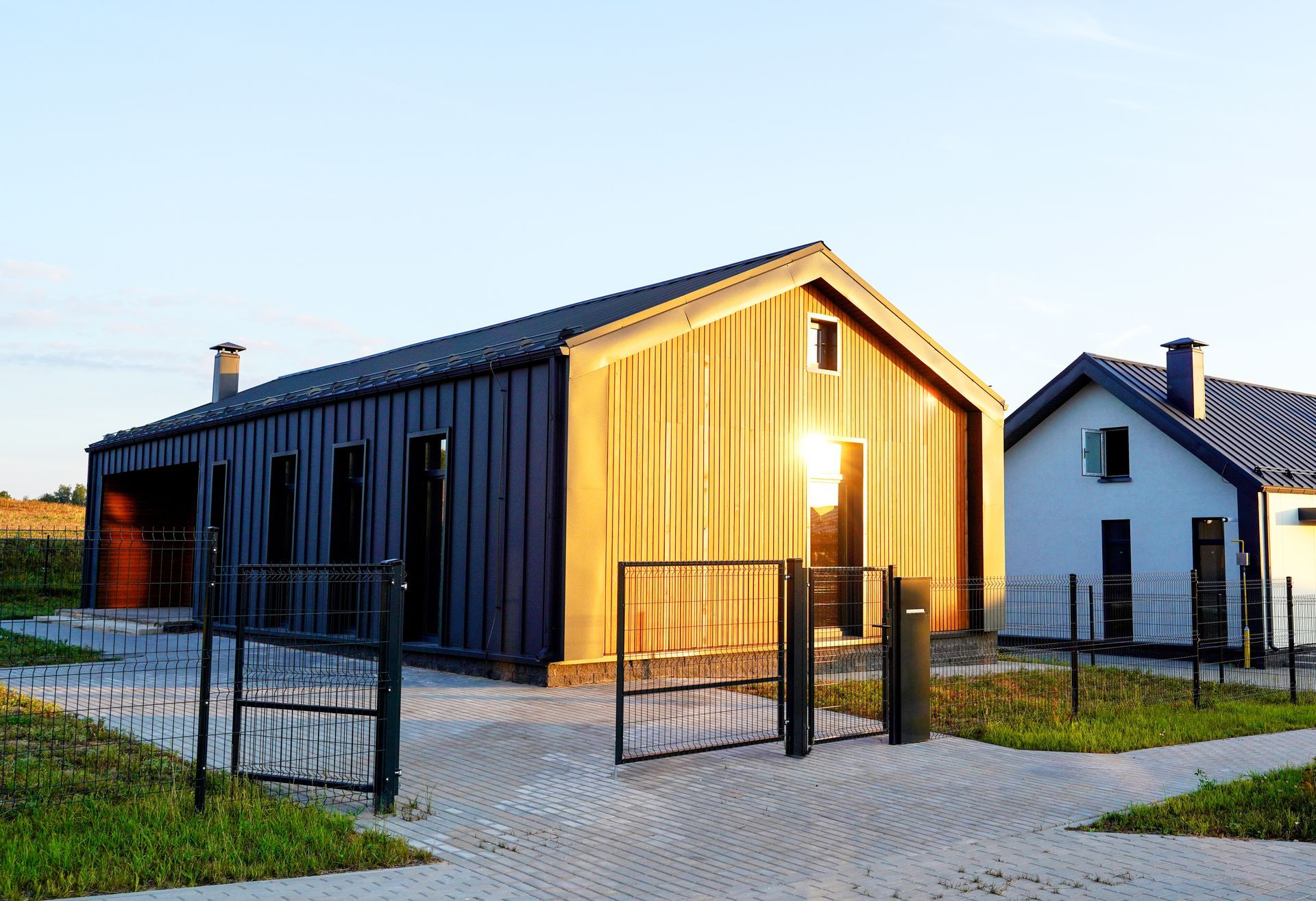 Two modern houses with contrasting facades: one blue-gray, the other wood-toned; sunlit, surrounded by brick pathways.