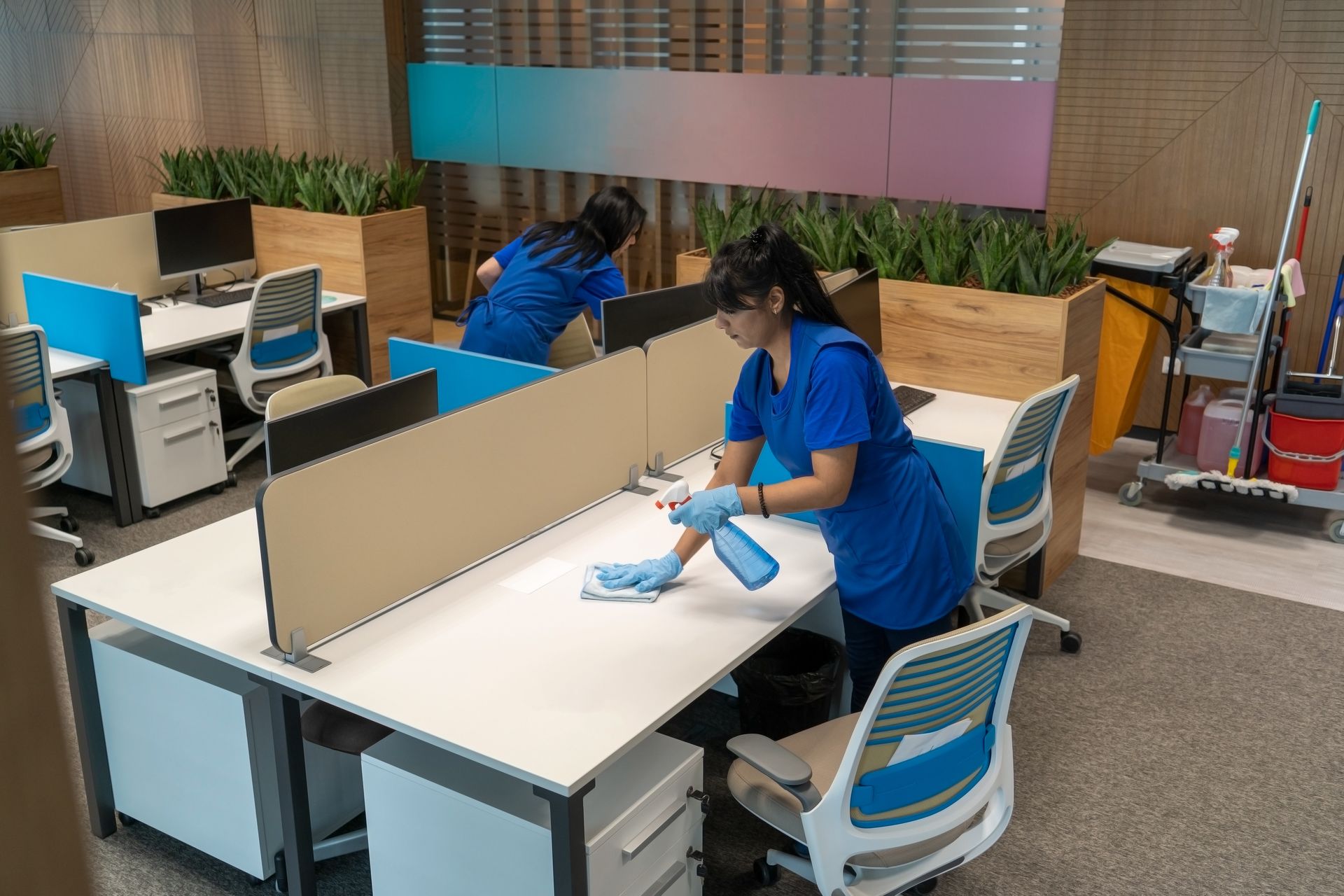 Two people in blue cleaning an office. One sprays and wipes a desk, the other cleans nearby.