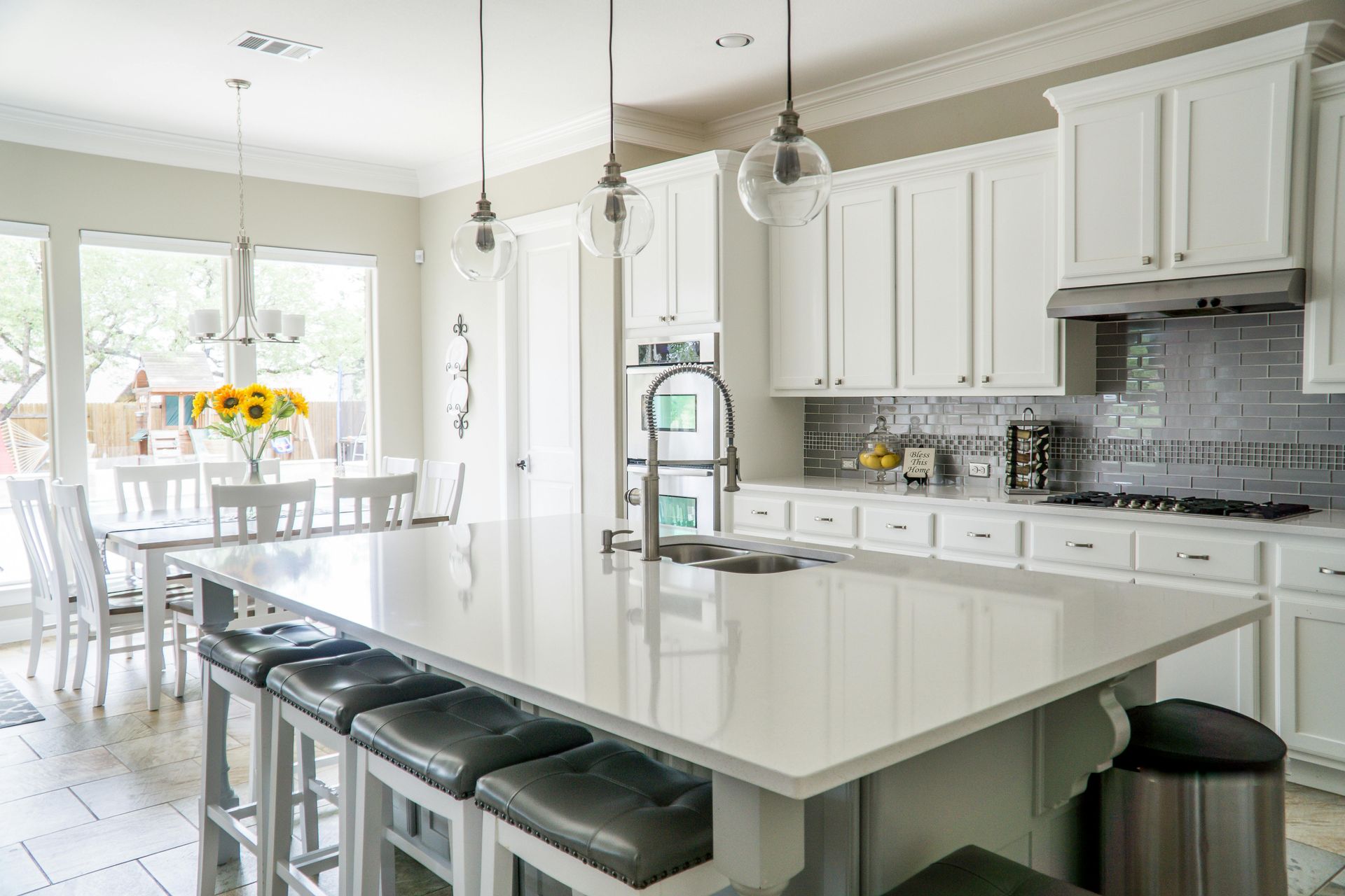 White kitchen with island, seating, and stainless steel appliances. Dining table visible with large windows.