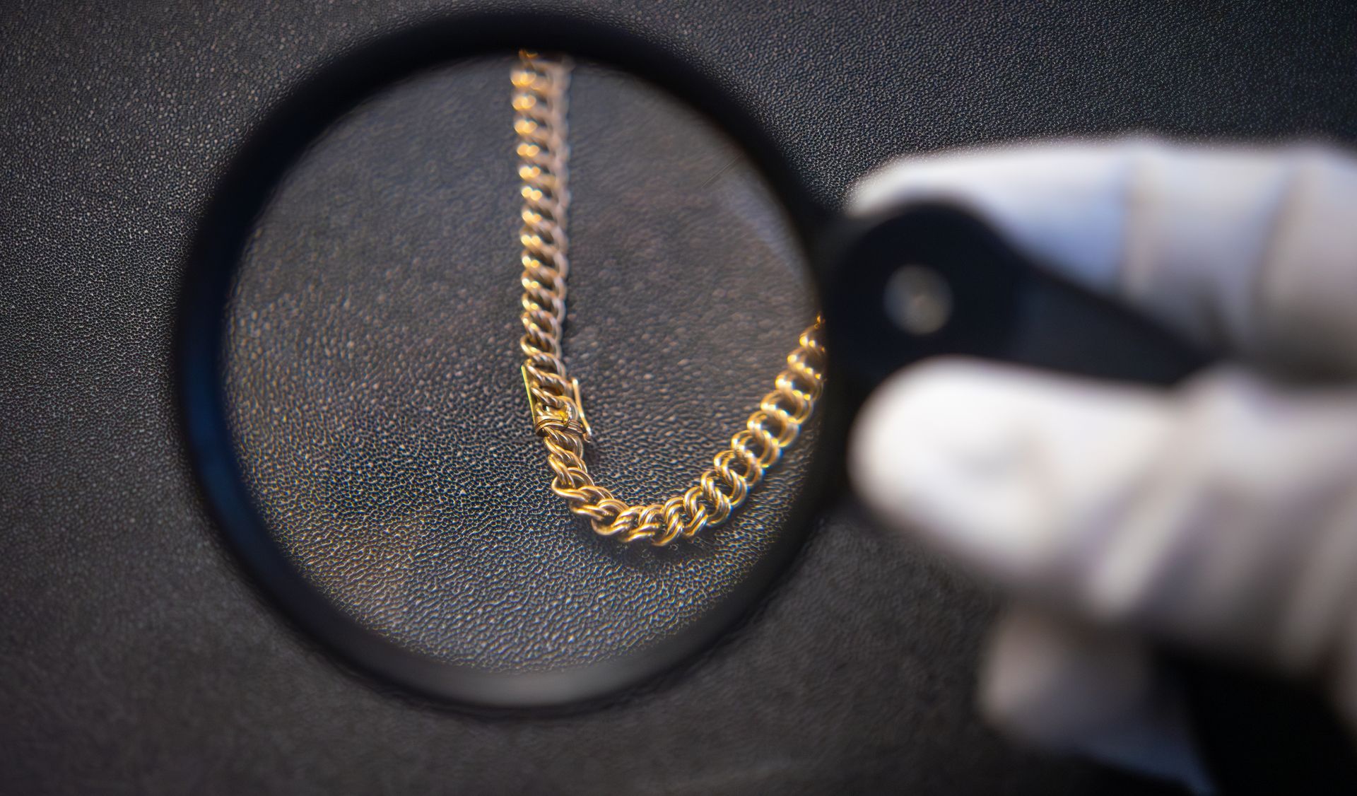 A gloved hand examining a gold chain bracelet through a magnifying loupe for appraisal