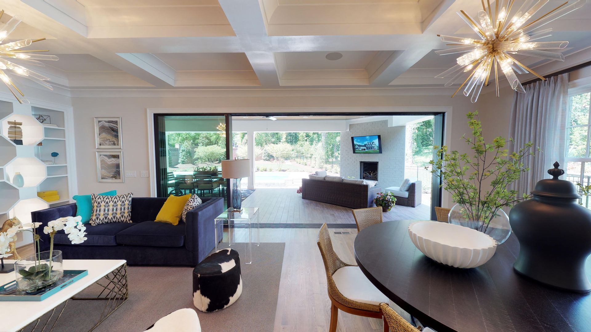 Modern open-plan living room with dark sofa, dining table, starburst lights, and large windows to a patio