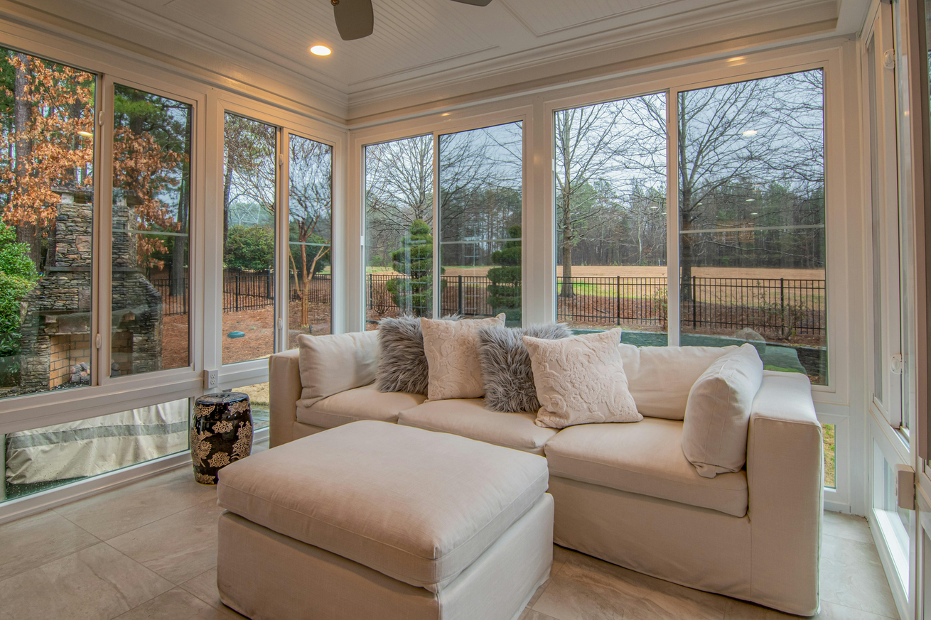 Bright sunroom with beige sofa and ottoman, surrounded by large windows overlooking a wooded yard.