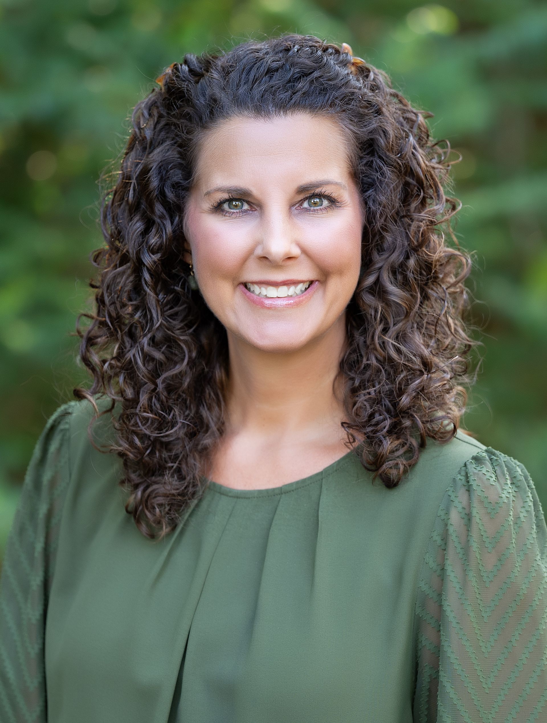 A woman with curly hair is wearing a green shirt and smiling for the camera.